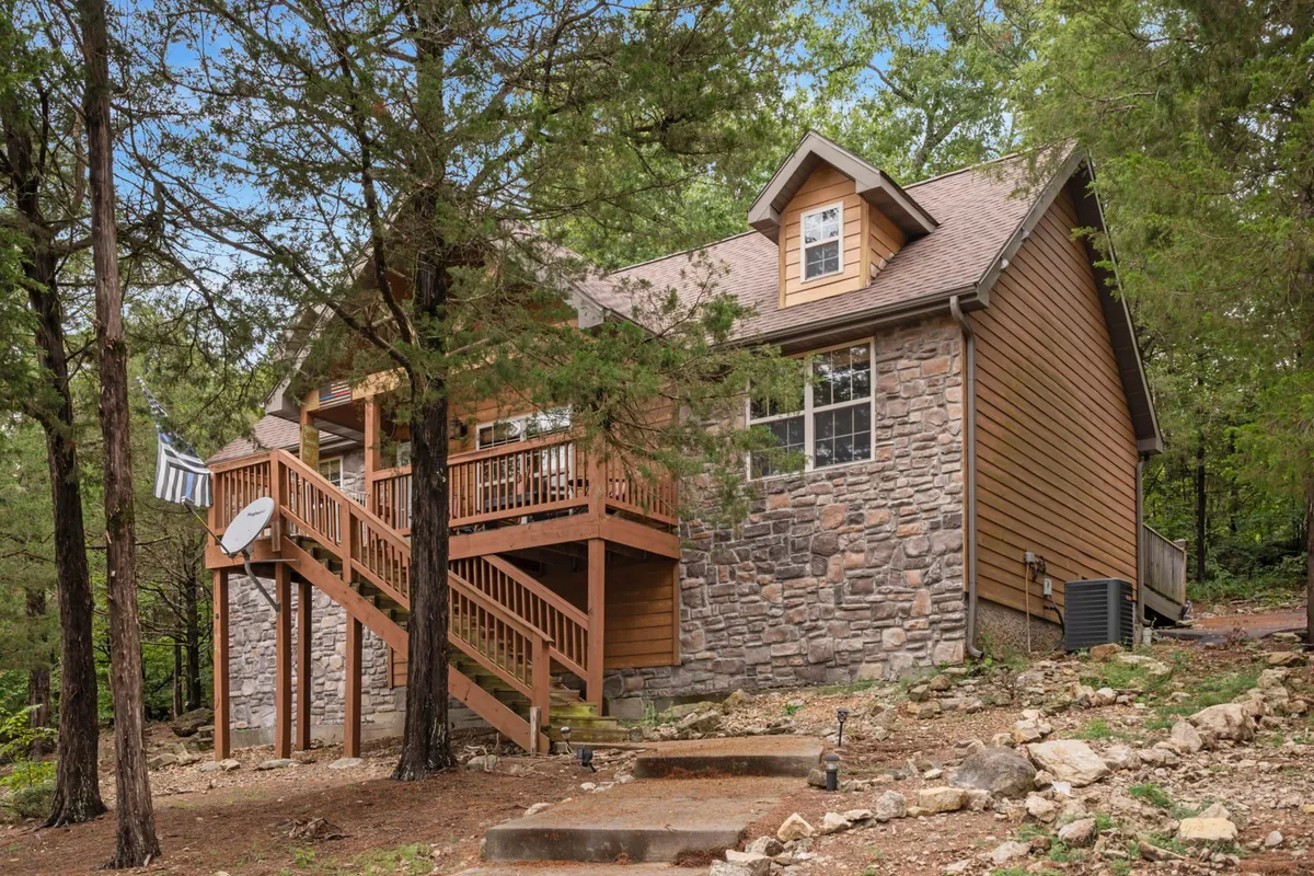 A peaceful exterior view of the lodge's wooden deck with a stone facade, staircase, and surrounding trees. The scene suggests a quiet, natural setting perfect for relaxing or gathering outdoors, with the forested surroundings visible.