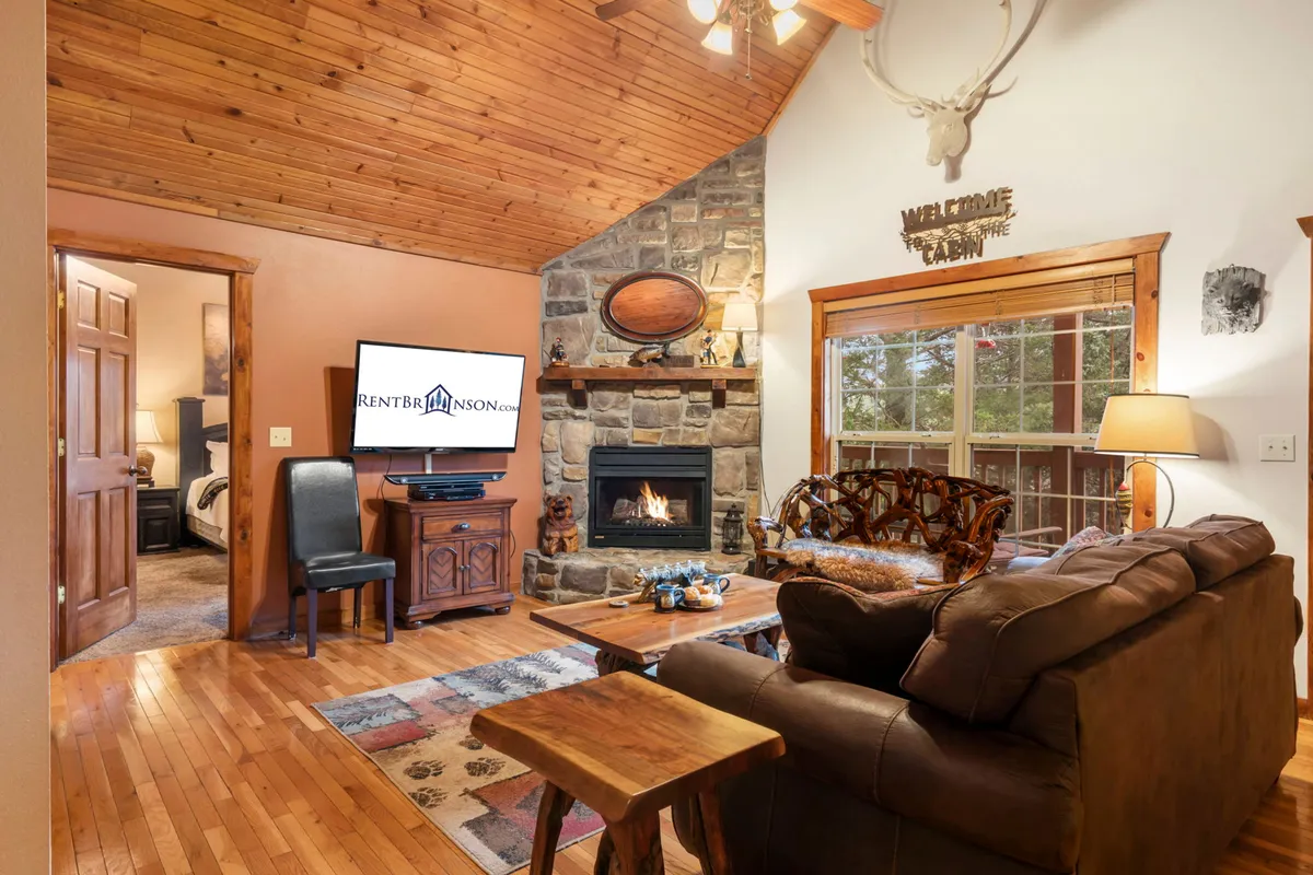 The image showcases a cozy lodge interior featuring a striking stone fireplace, vaulted wooden ceilings, plush seating, and a television—creating an inviting atmosphere perfect for relaxation or social gatherings.