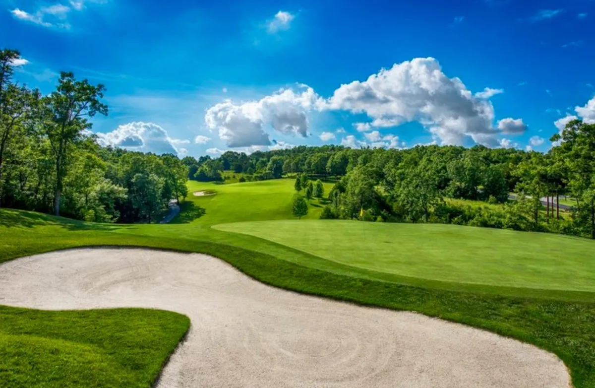 A scenic shot of the lush green fairway of a championship golf course, with a sand bunker, rolling hills, and a blue sky overhead. The landscape feels open and inviting, ideal for resort activities like golfing or nature walks.