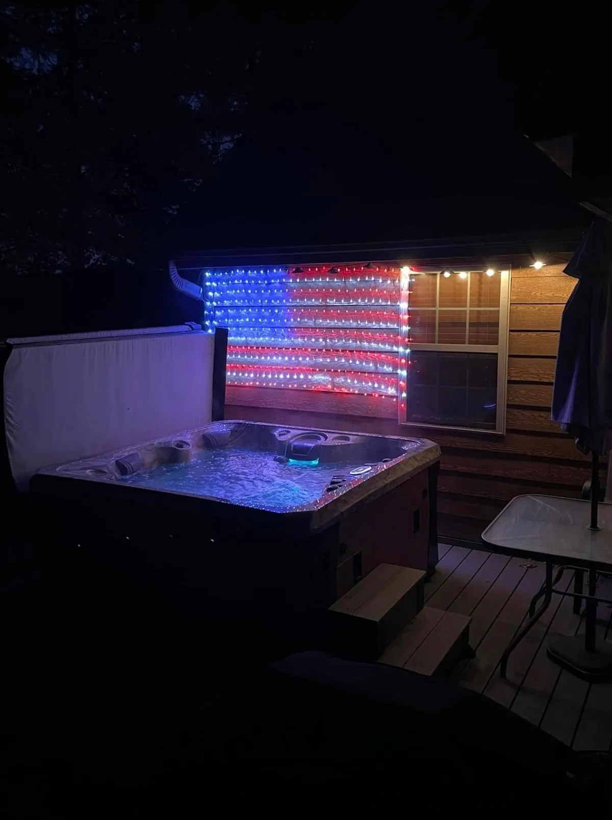 A bubbling hot tub on a wooden deck is accented by string lights and comfortable patio furniture, inviting guests to unwind outdoors. American flag lights add a festive touch, hinting at memorable evenings under the stars.