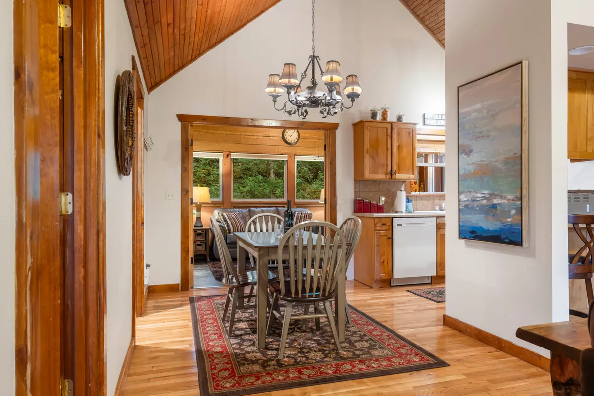 A bright and inviting dining space featuring a large dining table beneath a beautiful chandelier, with expansive windows letting in natural light. The kitchen cabinetry and a patterned rug add warmth, making it ideal for both casual meals and special celebrations.