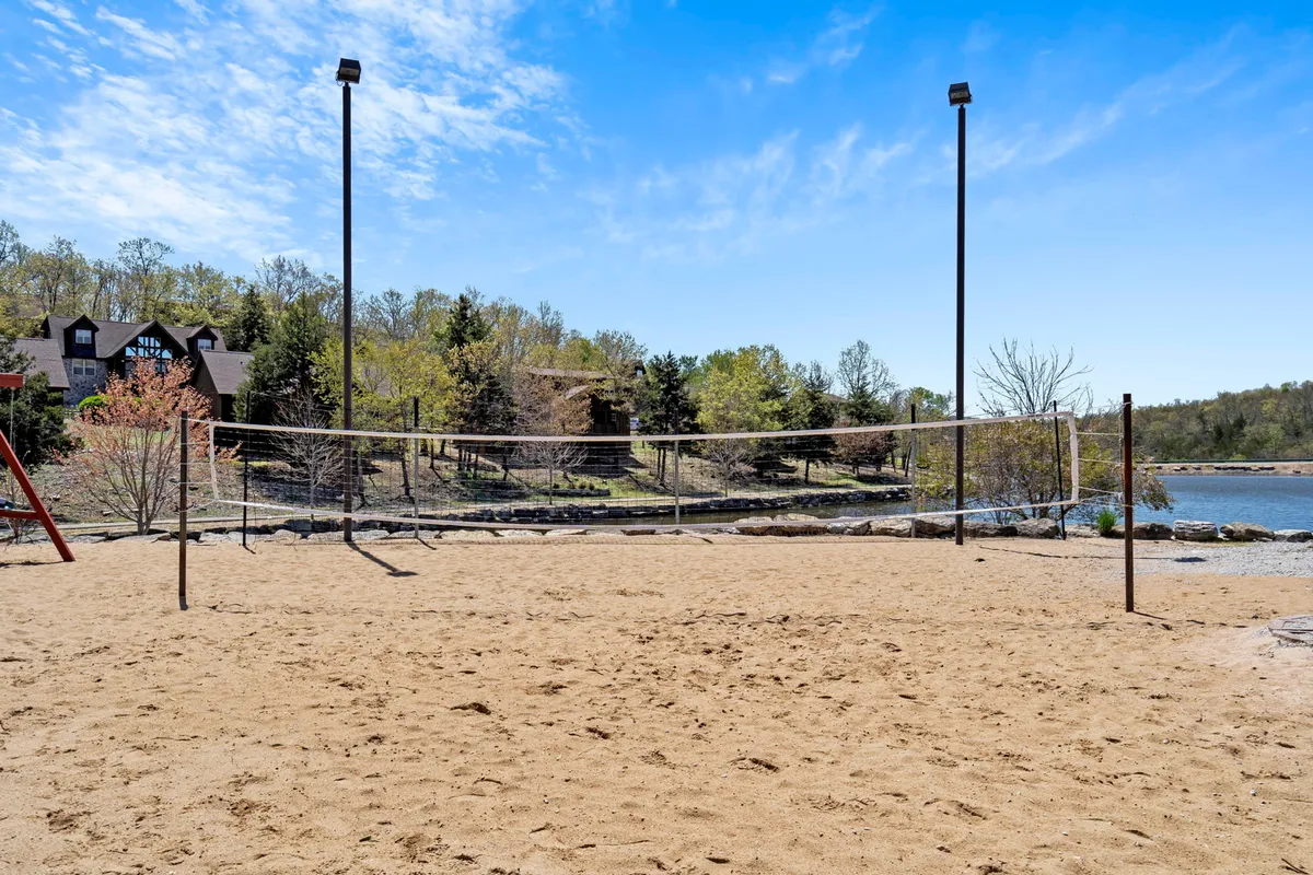 An active sand volleyball court sits near a scenic lake, with trees and a lodge in the background. The space is alive with the possibility of friendly games and group activities in a cheerful, natural setting.