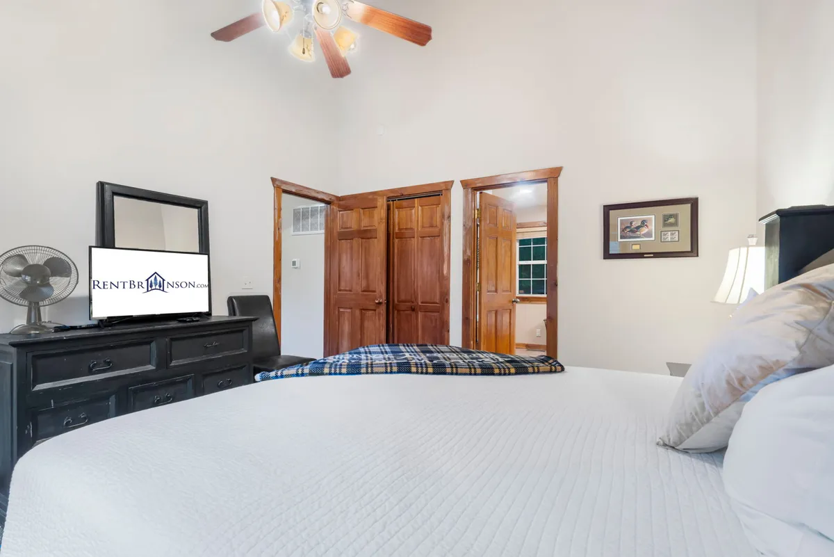 This image features a cozy bedroom with a king-sized bed dressed in crisp white bedding, a wall-mounted television for private entertainment, a wooden dresser, double doors leading to another room, and framed artwork. The room is inviting, with soft lighting and a tranquil atmosphere encouraging rest and relaxation.